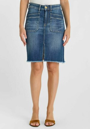 Jupe en denim avec poches plaquées à l'avant, fente frontale, ourlet effiloché, portée avec un haut blanc et des sandales ouvertes marron.