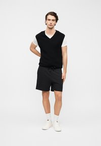 Μαύρο πλεκτό αμάνικο πουλόβερ πάνω από λευκό t-shirt, συνδυασμένο με μαύρο σορτς. Το μοντέλο φοράει λευκά αθλητικά παπούτσια και κάλτσες μέχρι τον αστράγαλο. Ουδέτερο φόντο.