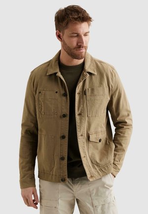 Man met baard in een beige button-up jas en beige cargobroek, staand met één hand in de zak, kijkend naar beneden en opzij.