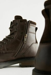 Botas de tobillo de cuero marrón con cordones en la parte delantera y cremallera lateral, con un cuello de ante texturizado y un tacón apilado.