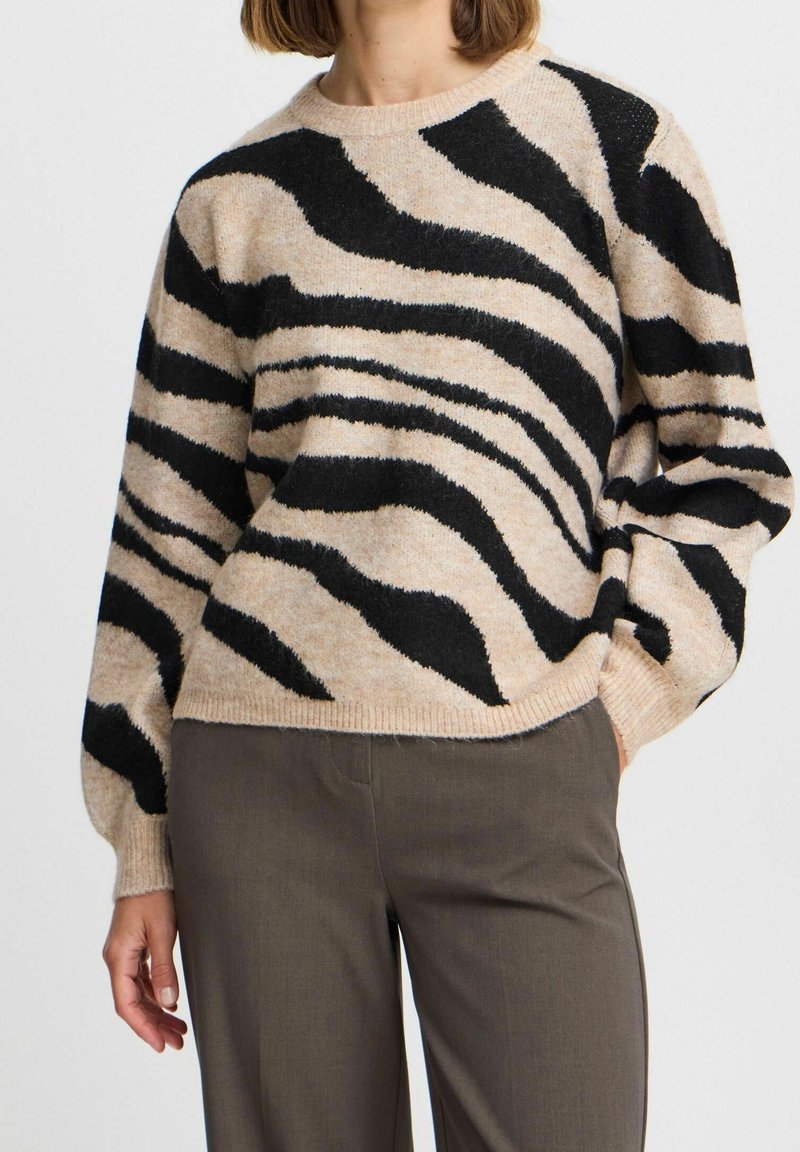Femme portant un pull beige avec des rayures zébrées noires et un pantalon marron, debout avec une main dans la poche devant un fond uni.