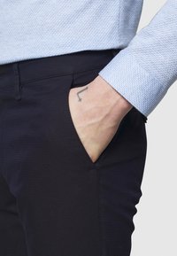 Jasnoniebieska, teksturowana koszula z wzorem w zygzak, noszona z czarnymi spodniami z boczną kieszenią. Ręka spoczywa w kieszeni, odsłaniając tatuaż.