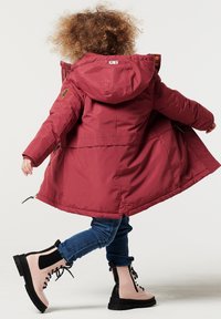 Chaqueta acolchada roja con capucha, cremallera y cierres a presión. Llevada sobre unos vaqueros azules y unas botas rosas con acentos en negro, que cuentan con una suela gruesa.