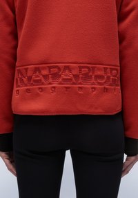 Giacca in pile rossa con logo in rilievo di un rosso più scuro sul bordo. I pantaloni neri indossati sotto la giacca creano un contrasto.