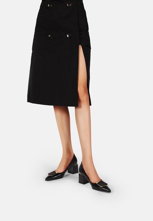 Femme portant une jupe noire mi-longue avec une fente sur le côté et des chaussures à talons bas noires avec des boucles décoratives.