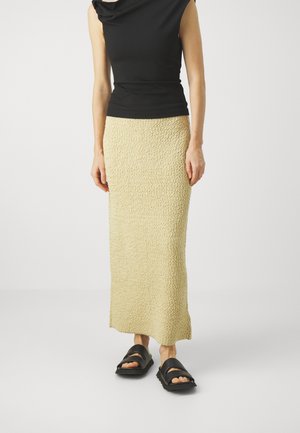 Top noir sans manches avec un décolleté drapé, associé à une jupe maxi beige texturée et des sandales noires à larges brides et semelles épaisses.