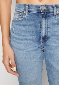 Jeans de mezclilla de color azul claro con cintura alta, detalles desgastados y herrajes en tono cobre. Presenta bolsillos delanteros y cierre de cremallera.