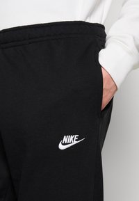 Närbild av en persons hand i fickan på svarta Nike sweatpants med vit logga, iförd en vit långärmad tröja mot en enkel bakgrund.