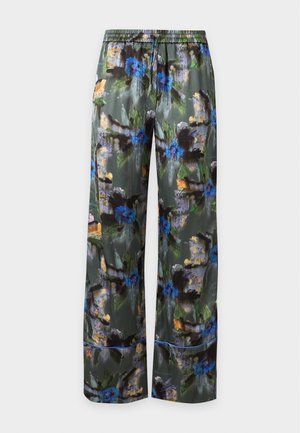 Femme portant un crop top blanc sans manches et un pantalon ample vert à imprimé floral avec des sandales noires à talons hauts, debout les mains dans les poches.