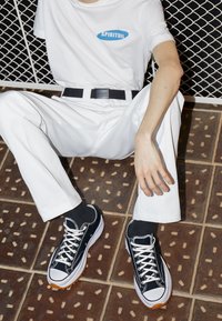 Baskets en toile noire avec des lacets blancs et des semelles en caoutchouc, portées avec un pantalon blanc et une chemise blanche avec un logo bleu. Fond en carrelage texturé.