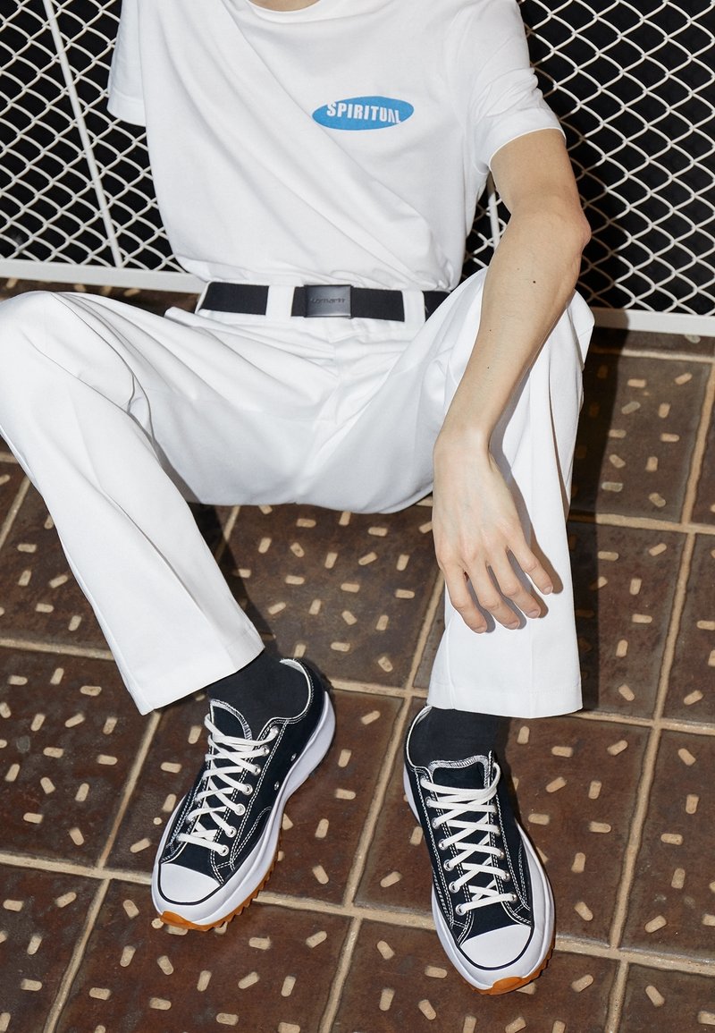 Baskets en toile noire avec des lacets blancs et des semelles en caoutchouc, portées avec un pantalon blanc et une chemise blanche avec un logo bleu. Fond en carrelage texturé.
