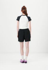 Tinkama balta palaidinė su juodais raglano rankovėmis, derinama su juodomis aukšto liemens šortais. Avalynė purpurinė su baltais akcentais; modelis stovi nugarą pasukęs.