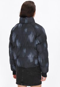 Pufferjack met hoge kraag, zwart en blauw tie-dye patroon, met gequiltte secties en geribde manchetten. Gecombineerd met een zwarte denim rok.