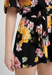 Kukkakuvioinen haalari, jossa on musta tausta vaaleanpunaisilla ja keltaisilla ruusuilla, joustava vyötärö ja sidont yksityiskohta vyötäröllä. Pehmeä kangaspinta.