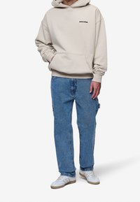 Sudadera beige con capucha y bolsillo frontal, logo negro; combinada con pantalones cargo de mezclilla azul claro; zapatillas blancas con toques de marrón.