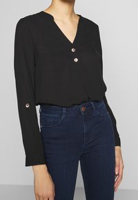 Blouse noire à manches longues avec décolleté en V, comportant deux boutons et des accents dorés décoratifs. Le tissu a une texture lisse. Portée avec un jean bleu.
