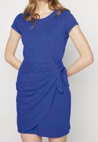 Modrá šaty s krátkým rukávem vyrobené z měkké látky, s nařasenou stranou a uvázaným akcentem, a skromným kulatým výstřihem.