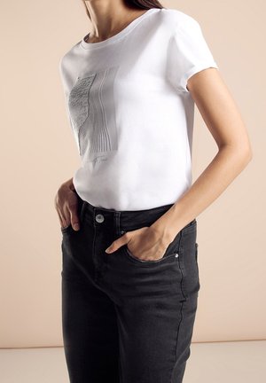 Woman wearing a white graphic t-shirt with abstract black and gray lines, tucked into high-waisted black jeans, hands in pockets.