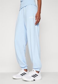 Person som bär ljusblå Adidas träningsbyxor och vita Adidas sneakers med svarta ränder, stående mot en neutral bakgrund.