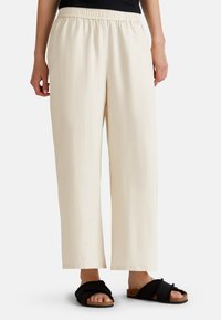 Pantalones elásticos de talle alto en beige claro, con un corte holgado y piernas rectas. Combinados con sandalias negras y peludas con suela de corcho y bandas cruzadas.