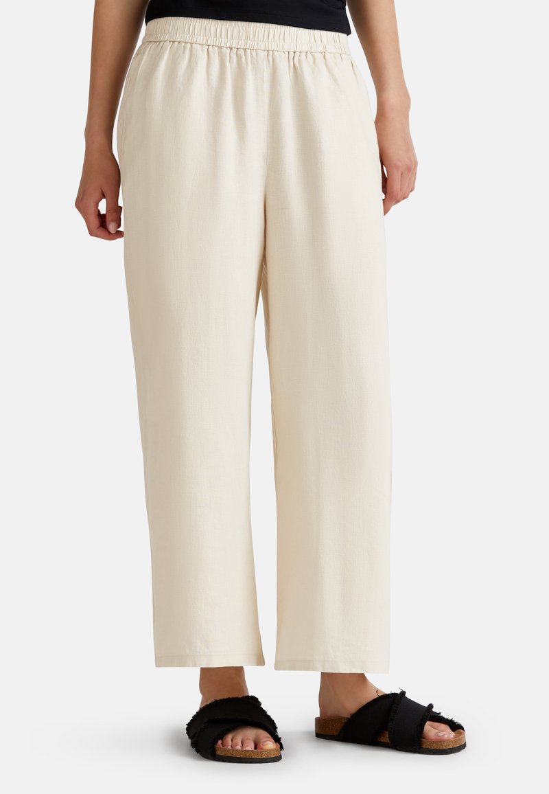Pantalones elásticos de talle alto en beige claro, con un corte holgado y piernas rectas. Combinados con sandalias negras y peludas con suela de corcho y bandas cruzadas.