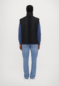 Black patterned vest with a high neck, featuring a textured back and ribbed blue long sleeves, paired with light blue jeans.