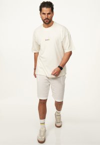 Ecrufarbenes Baumwoll-T-Shirt mit kurzen Ärmeln und kleinen mehrfarbigen Designs auf der Vorderseite, kombiniert mit hellbeigen, strukturierten Shorts und Sneakern.