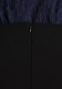 Vestido negro con un cuerpo de encaje con patrón floral y una cremallera oculta en la parte posterior. La tela suave se extiende debajo de la cintura.