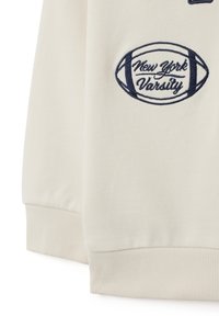Sweat-shirt couleur crème avec des poignets et un ourlet côtelés. Présente une broderie marine d'un football et le texte "New York Varsity". Texture douce.