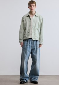 Τζιν μπουφάν ανοιχτού πράσινου χρώματος με κουμπιά μπροστά, δύο τσέπες και άνετη εφαρμογή, συνδυασμένο με υπερμεγέθη μπλε τζιν παντελόνια. Μαύρα παπούτσια ολοκληρώνουν την εμφάνιση.