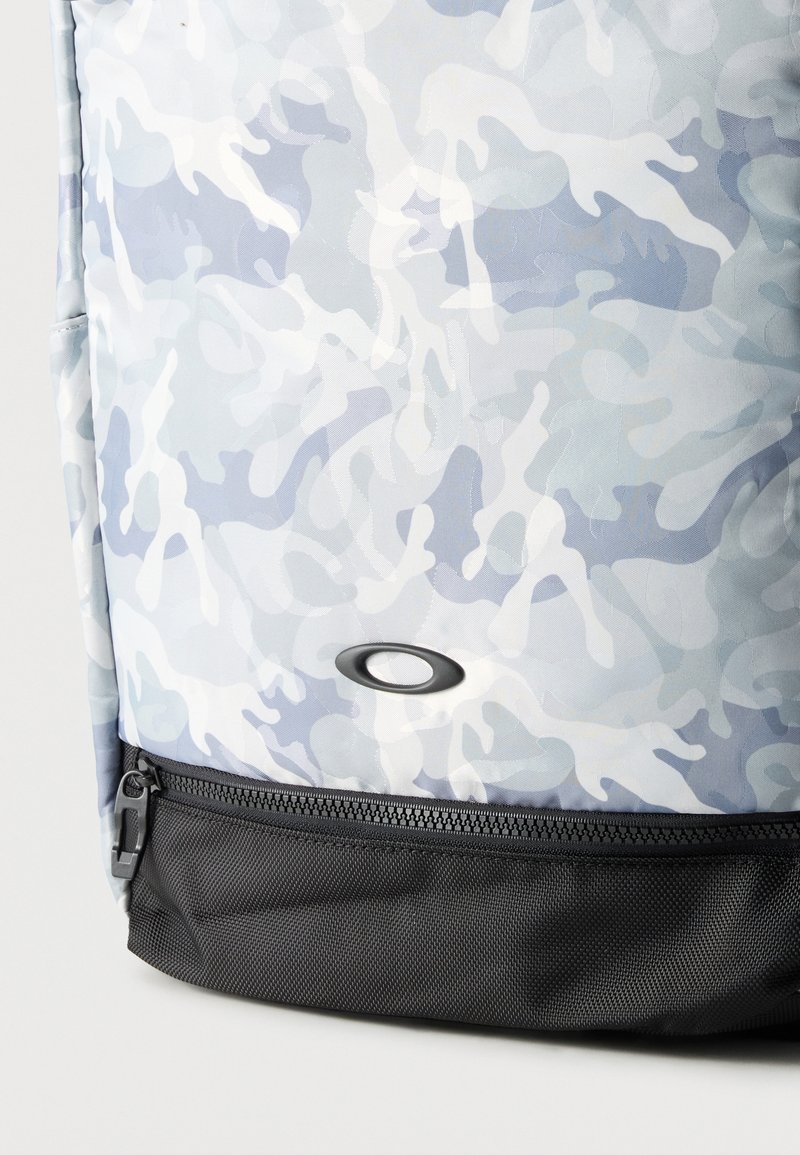 Lyseblå og hvide camouflage rygsæk med sort lynlåslomme i bunden og oval sort logo.
