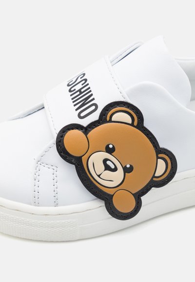 Hvit syntetisk sneakers med en fremtredende brun bjørn-applikasjon og svarte detaljer, med borrelåsrem og teksturert såle.