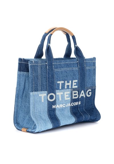 Džinsinis pirkinių krepšys su įvairiais mėlynai atspalviais, ryškiu baltu tekstu, kuriame užrašyta "THE TOTE BAG MARC JACOBS", sustiprintomis rankenomis, struktūruota forma.