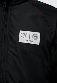 Sort sportsjakke med lynlås, røde detaljer og et hvidt mærke med teksten "HALO" og "OFF PITCH". Glat stofstruktur.