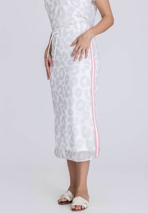 Femme portant une jupe midi blanche à imprimé léopard avec des bandes latérales roses et des sandales blanches, debout devant un fond clair uni.