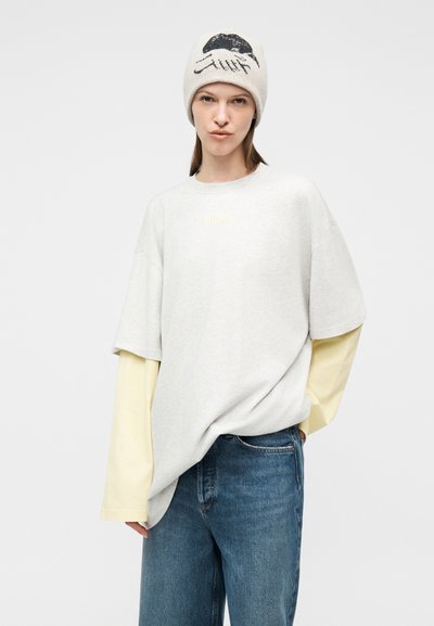 Νεαρό άτομο που φοράει λευκό φαρδύ T-shirt πάνω από ανοιχτό κίτρινο μπλουζάκι με μακριά μανίκια, μπλε τζιν και γκρι πλεκτό καπέλο με μαύρο και άσπρο σχέδιο.