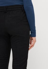 Pantalones de pana negra con una superficie texturizada, que cuentan con una cintura elástica con cordón y bolsillos en la parte trasera. Una mano reposa sobre la cadera.