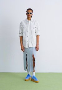 Chemise blanche à rayures, jupe fendue bleu clair, sneakers bleues avec semelles en caoutchouc, portant des lunettes de soleil blanches. Design minimaliste avec une coupe décontractée.