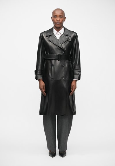 Manteau en cuir noir à double boutonnage avec une taille ceinturée et de larges revers, associé à un pantalon à motifs gris et des chaussures noires pointues.
