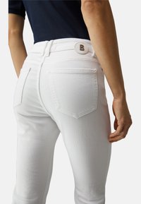 Jean en denim blanc avec une coupe ajustée, deux poches arrière, et un bouton rond marqué d'un 'B'. Texture lisse et coupe sur mesure visibles.