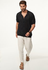 Schwarzes, offen gestricktes Kurzarmhemd mit Knopfleiste und aufwändigen Mustern, kombiniert mit weißen Leinenhosen und braunen Sandalen.