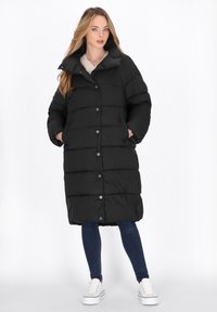 Sort puffer-jakke med en lang silhuet, kviltet design, trykknapper og sidelommer, båret over blå jeans og hvide sneakers.