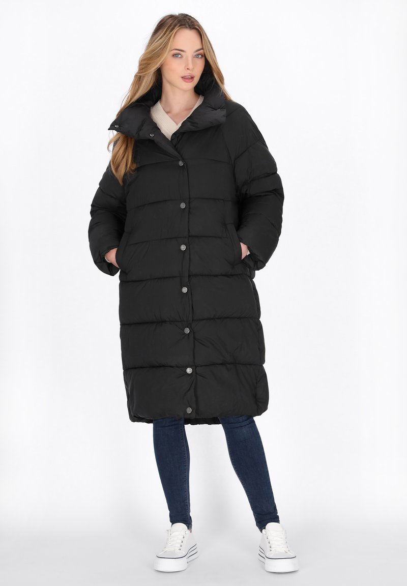 Sort puffer-jakke med en lang silhuet, kviltet design, trykknapper og sidelommer, båret over blå jeans og hvide sneakers.
