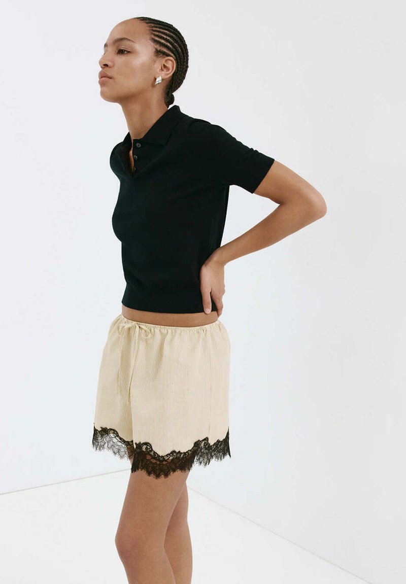 Femme aux cheveux tressés, debout avec une main dans le dos, portant un polo noir à manches courtes et un short beige avec bordure en dentelle noire.