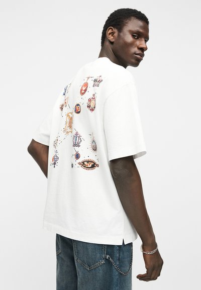 Uomo che indossa una T-shirt bianca oversize con stampe di emblemi colorati sul retro, abbinata a jeans di denim blu e un braccialetto a catena d'argento.