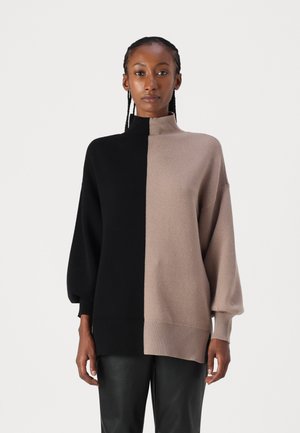 Pull oversized avec col montant ; présente des sections moitié noires, moitié beiges, des poignets et un ourlet côtelés, texture en maille lisse.