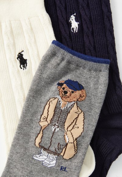 Trois paires de chaussettes : une paire en maille torsadée blanche, une paire en côtes bleu marine et une paire grise avec un motif de ours. Chacune dispose de logos brodés, de textures variées et de couleurs différentes.