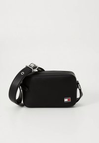 Schwarze Leder-Umhängetasche mit rechteckiger Form, silberner Hardware, Reißverschluss und einem kleinen Logopatch mit roten und blauen Streifen.