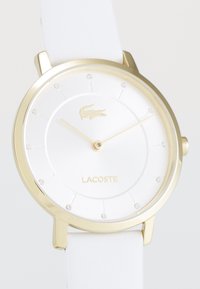 Lacoste-klocka med en rund vit urtavla, guldtonade visare, accentstenar och ett vitt läderarmband. Märkbar detalj med guld-logotyp.