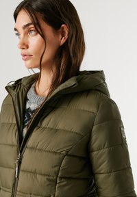 Olivza zaļa pūstošā jaka ar horizontālām šuvēm, pievienotu kapuci un priekšējo rāvējslēdzēju. Apakšā valkāta teksturēta vilnas džemperis ar tumšu rakstu.